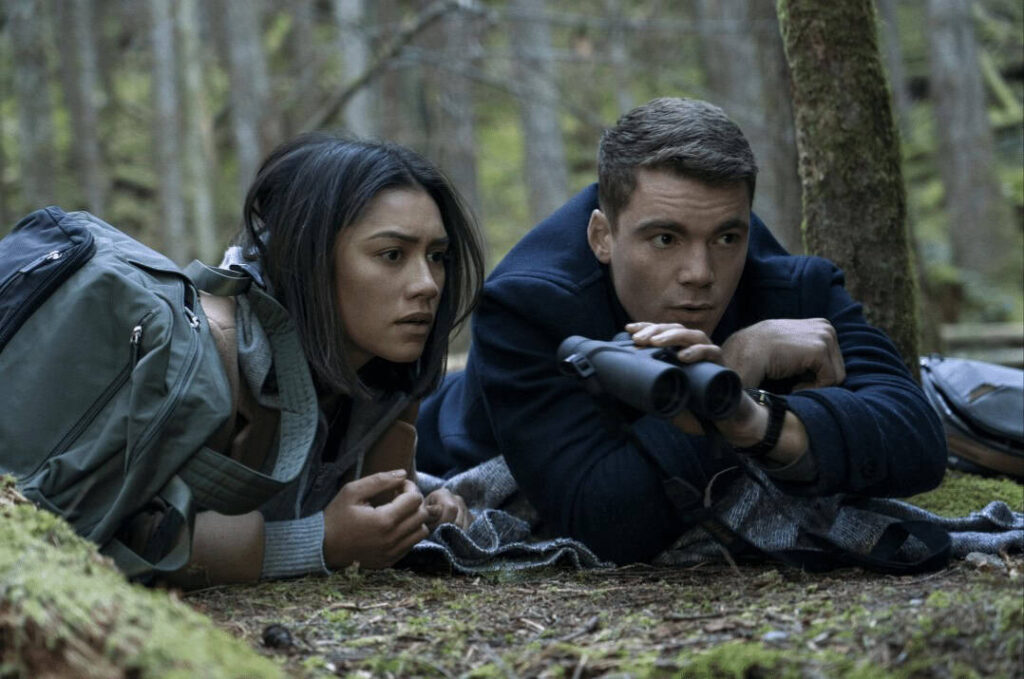 A tense forest scene from the Netflix show The Night Agent featuring two characters hiding and observing with binoculars.
