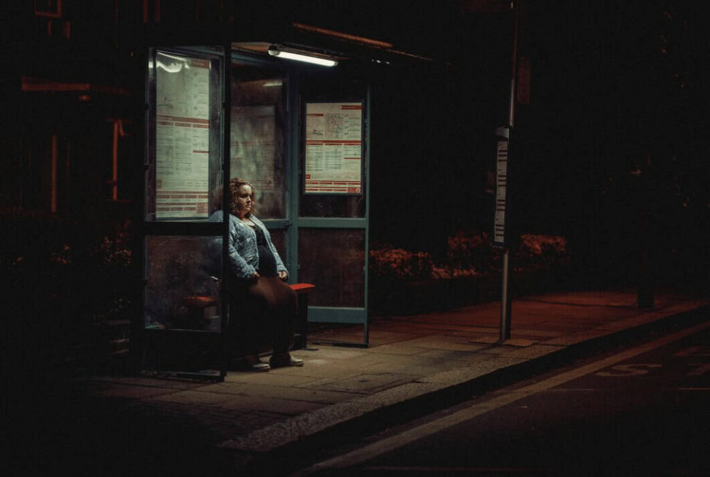 A lonely woman waits at a dimly lit bus stop in a tense scene from the Netflix show Baby Reindeer.