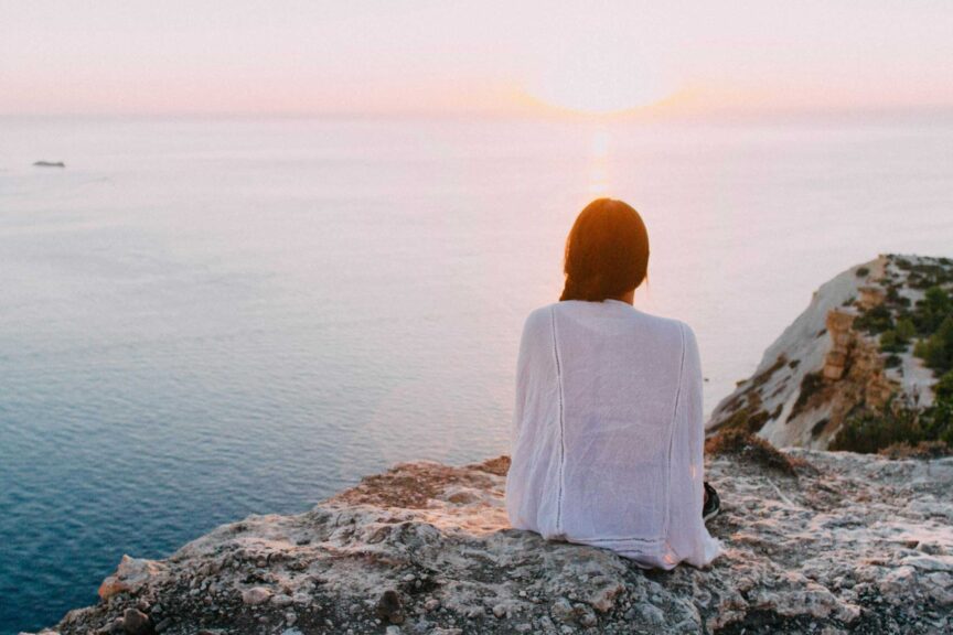 A serene view of a woman sitting on a rocky cliff, gazing at the ocean during sunset. Perfect for relaxation and travel themes. Mental Health