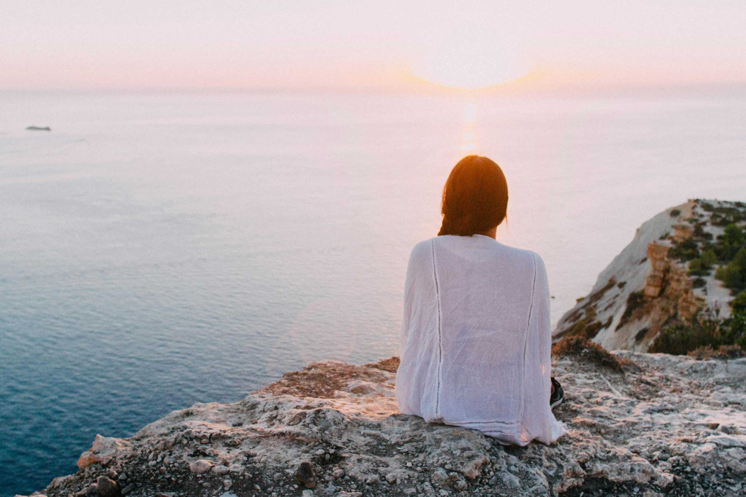 A serene view of a woman sitting on a rocky cliff, gazing at the ocean during sunset. Perfect for relaxation and travel themes. Mental Health