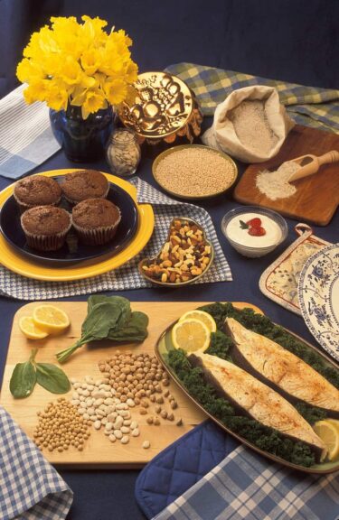 Plant-based foods rich in magnesium—bran muffins, pumpkin seeds, barley, buckwheat flour, yogurt, halibut, chickpeas, soy, spinach—arranged on a spring-themed presentation table with floral decor. Image by FotoshopTofs via Pixabay.