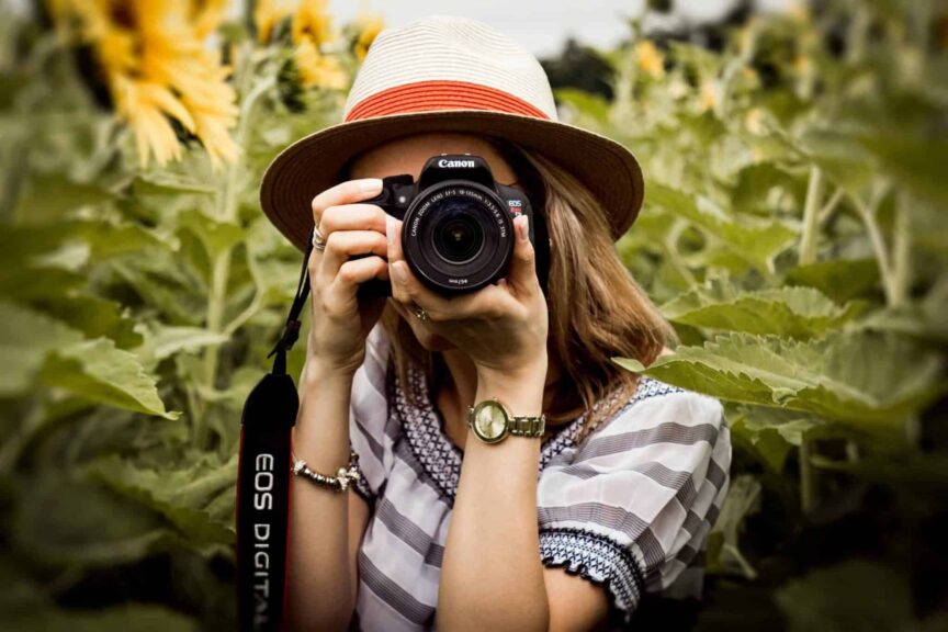 Woman with hat practicing travel photography in a summer sunflower field using a Canon camera. Image credit: by Andre Furtado via Pexels
