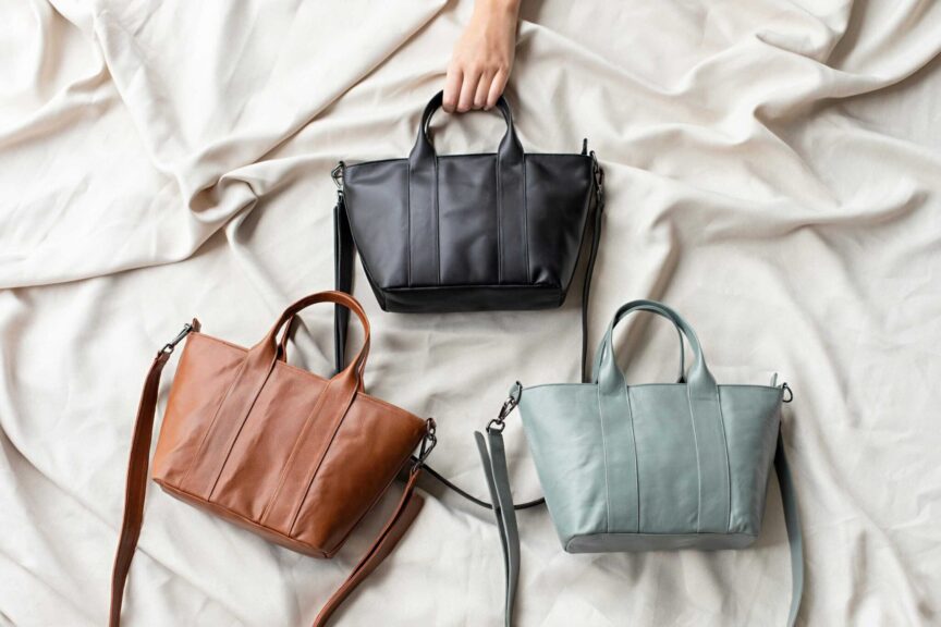 Top view of three stylish leather designer bags in black, brown, and gray on a soft fabric surface.