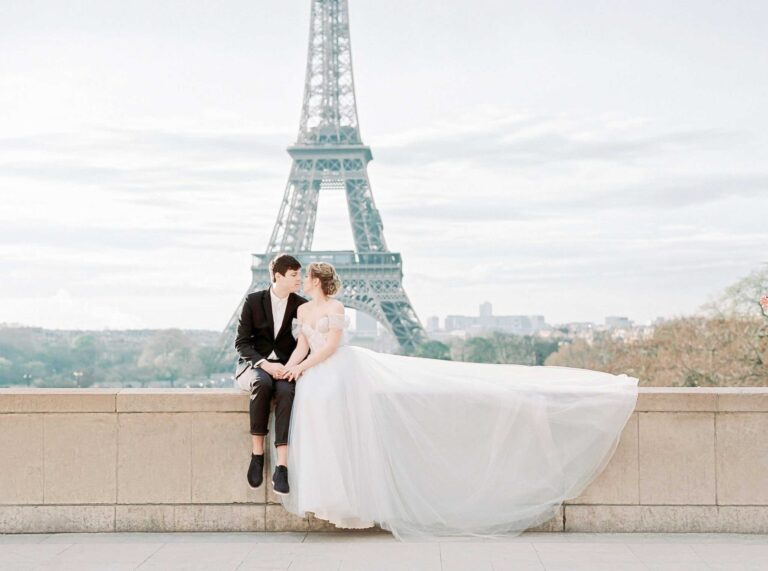 This photograph features a couple dressed for a wedding. They are sitting in front of the Eiffel Tower in Paris. Paris can be included in a list of honeymoon destinations.