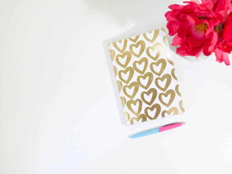 This photo features a journal with gold drawn hearts on the cover. There photo also contains flowers and a pen. Journaling can be a great way to develop a positive connection with yourself.