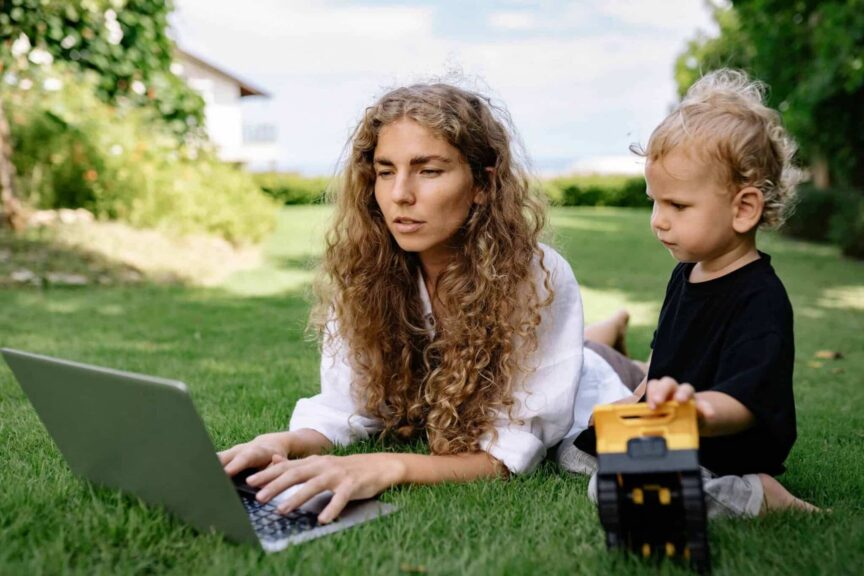 This photograph features a woman on a laptop. A kid is next to her with a toy. Parenting can include a balance of screen time and alternative activities.