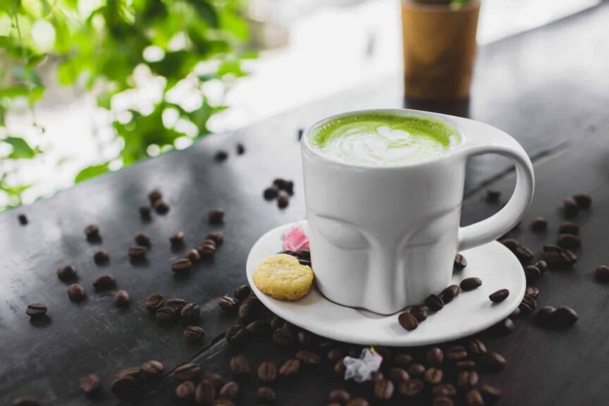 A vibrant matcha latte served with cookies on a saucer surrounded by coffee beans on a dark wooden surface.