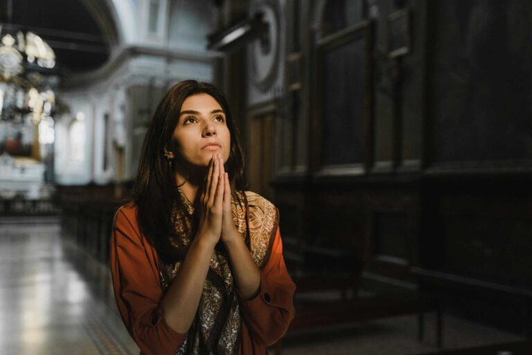 A woman is praying with hands clasped in a dimly lit church, creating a contemplative mood. Daily devotional