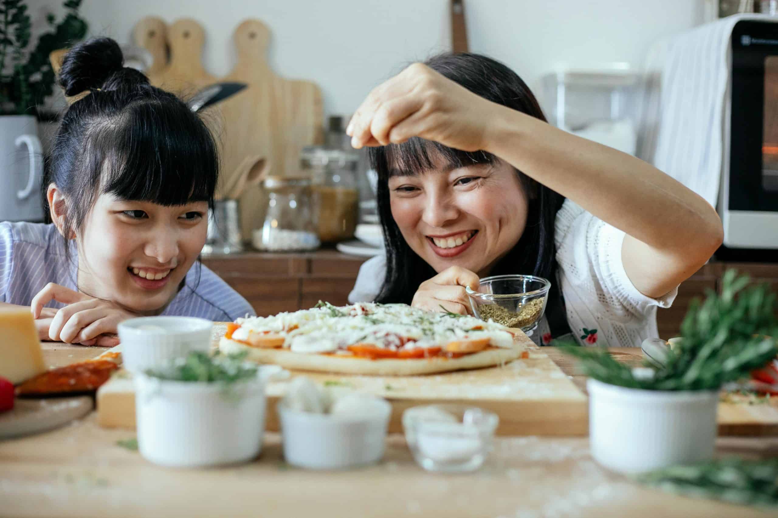 Two Asian women happily preparing a homemade pizza in a cozy kitchen setting.