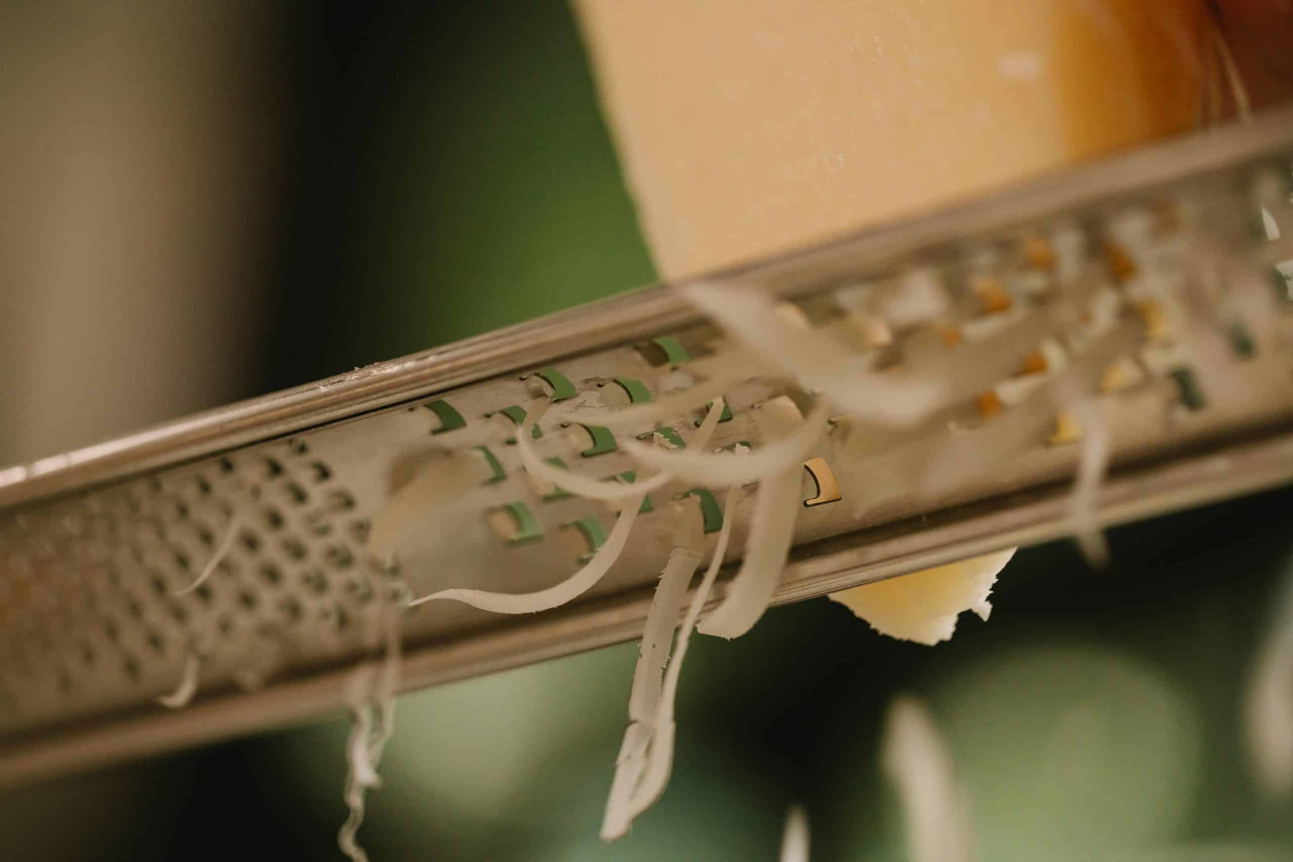 Closeup of tasty hard cheese grating on narrow stainless grater against blurred kitchen background