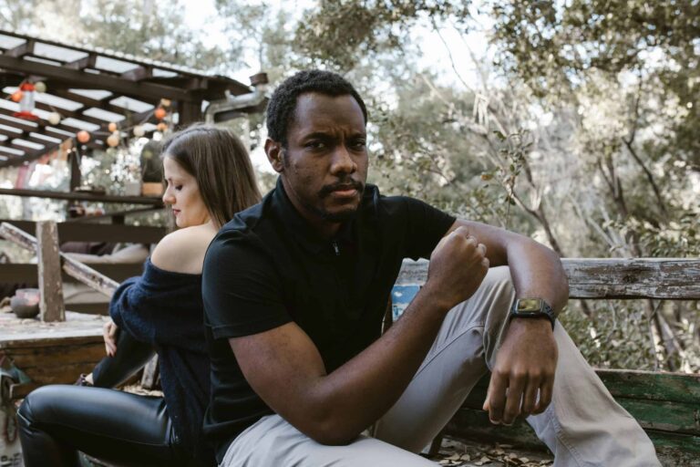 A thoughtful man sits outdoors, with a woman sitting distantly behind him. emotionally unavailable