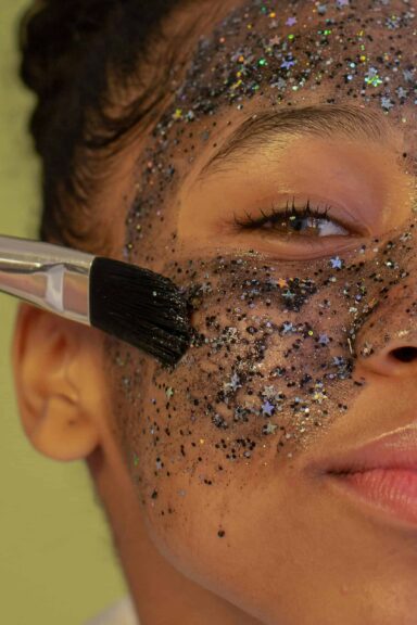 Crop unrecognizable young African American female with brush applying facial mask with glitters during beauty procedure and looking at camera on green background. Makeup hacks for Y2K