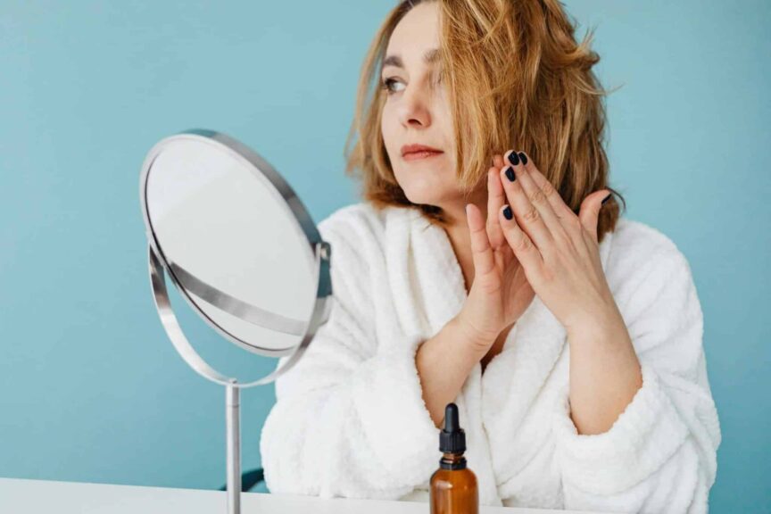 Woman in white robe applying skincare oil, looking in the mirror with manicured nails. beauty product launches