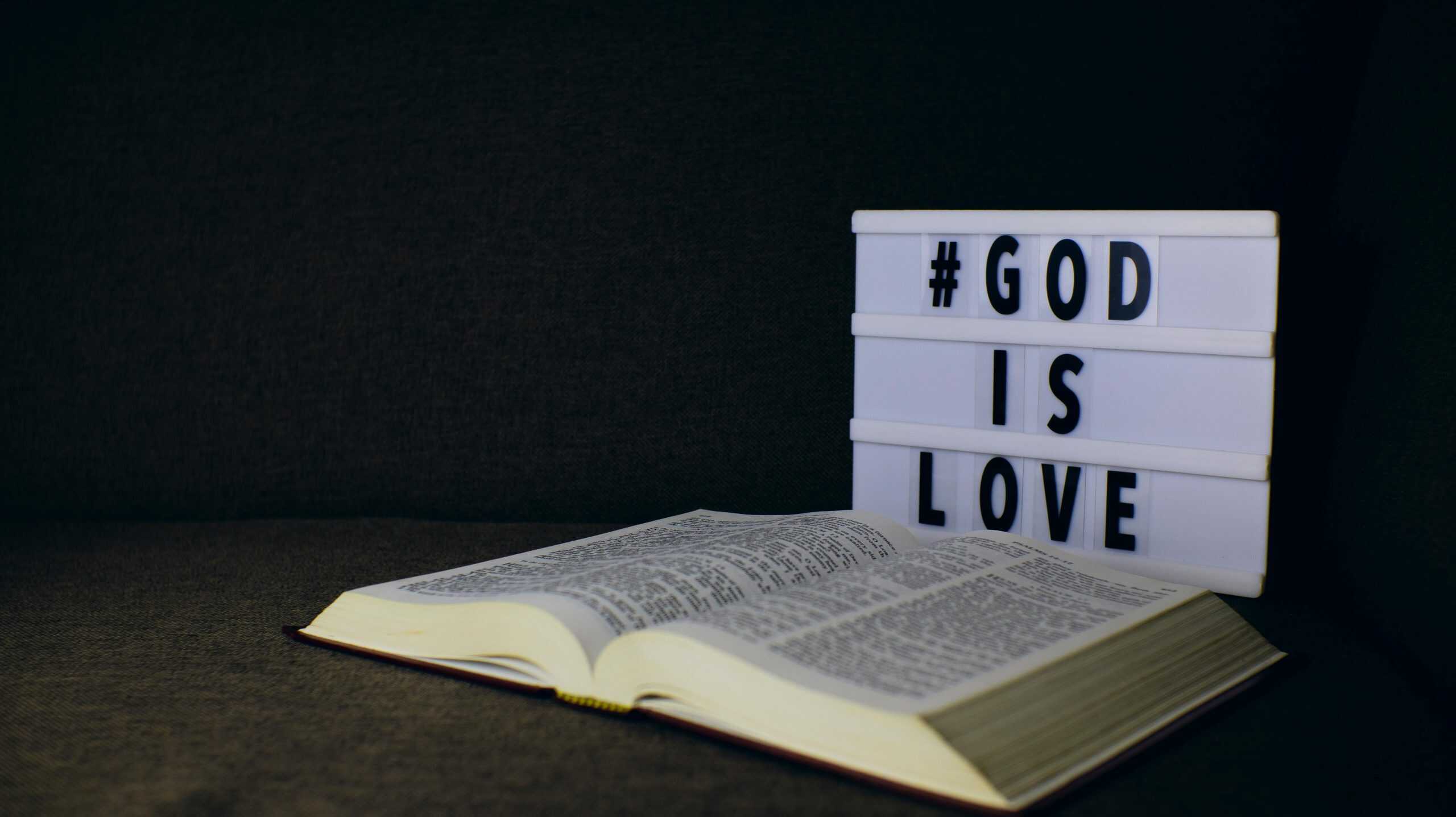 An open Bible next to a lightbox sign displaying the text 'God is Love' on a dark background. scripture, emotional maturity