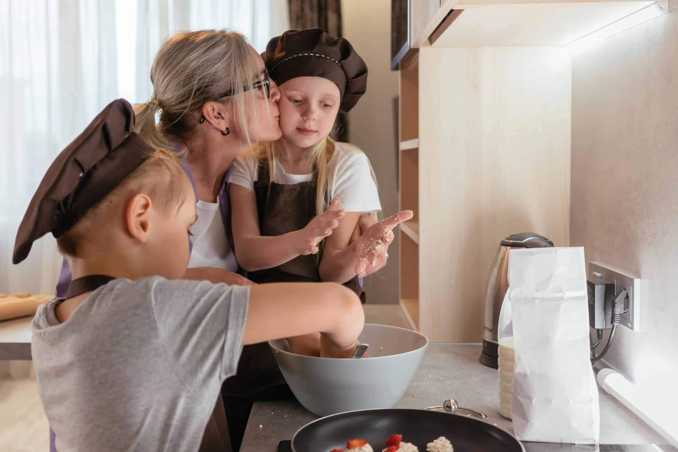 A loving grandmother kisses her granddaughter while baking with two children in the kitchen.