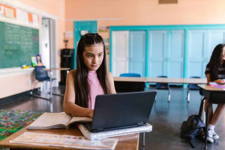 A focused young girl using a laptop for schoolwork in a bright classroom environment. back to school tech