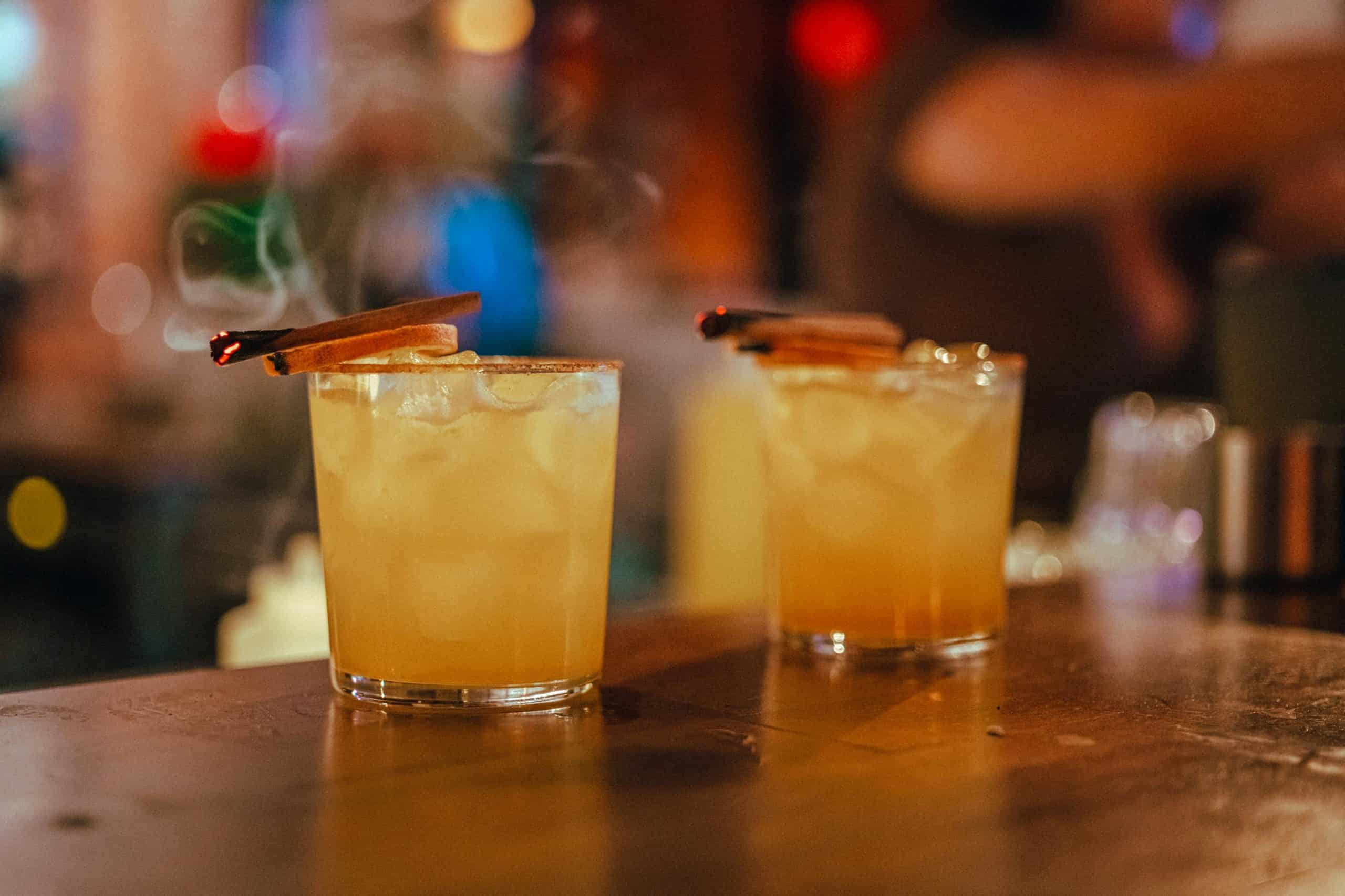 Two orange cocktails with cinnamon sticks on a wooden bar in a cozy, dim pub. Fall cocktail
