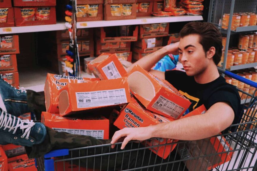 A man lounging in a shopping cart filled with Maruchan ramen in a supermarket aisle. ultra-processed foods