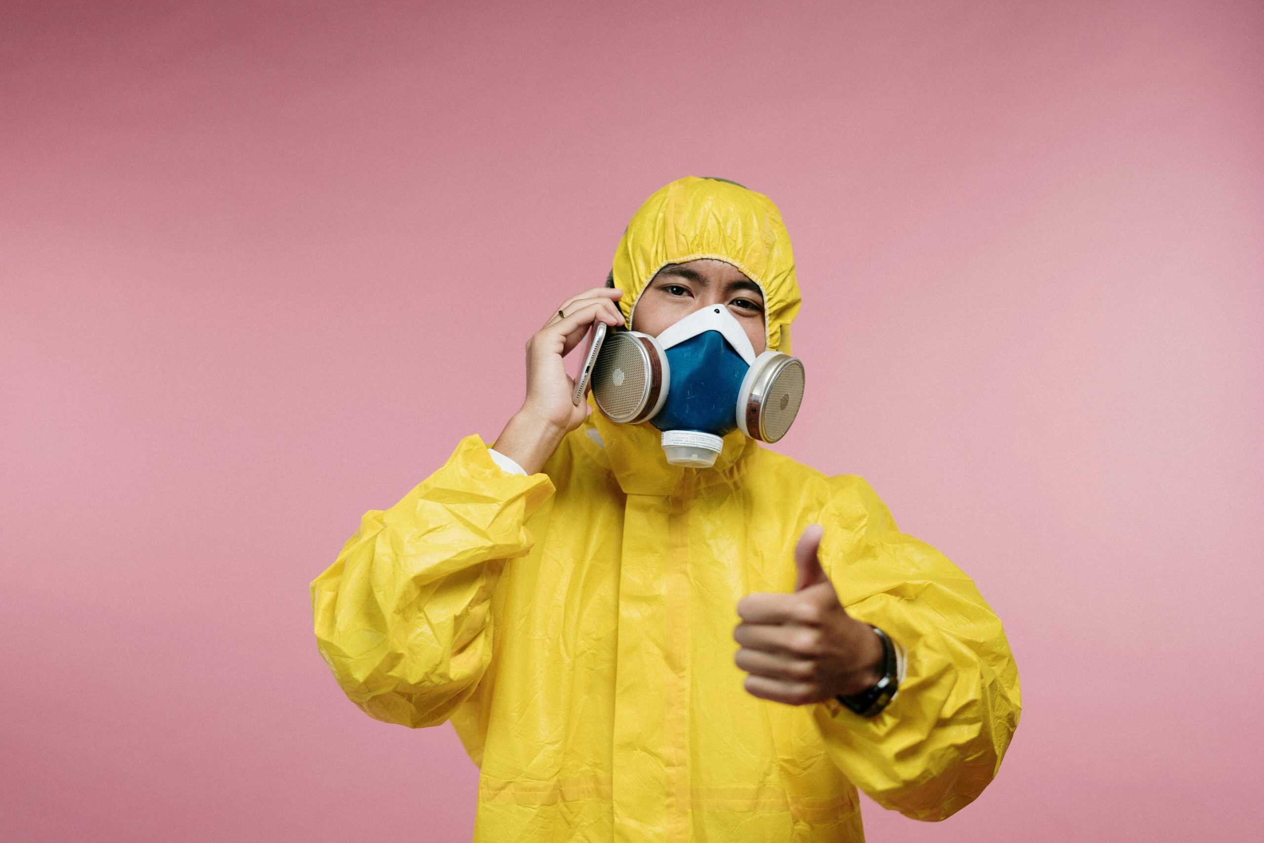 Individual in a yellow hazmat suit with a respirator giving a thumbs up, set against a pink background.