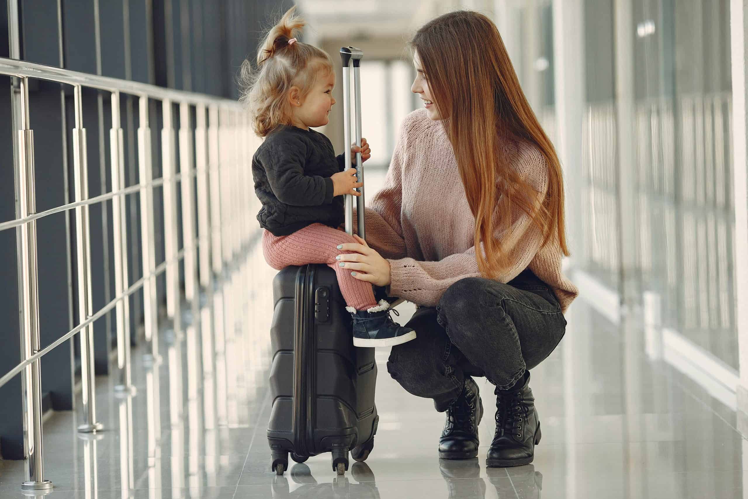 Full length of positive content woman and cute stylish blond little girl sitting on black suitcase in airport corridor while waiting for flight