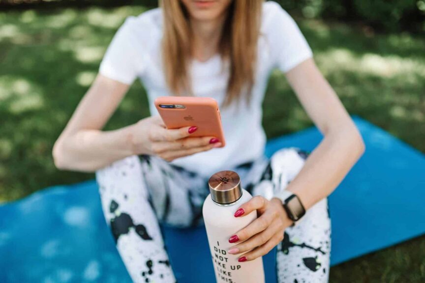 spirituality, technology, Woman using smartphone while holding bottle on yoga mat outdoors.