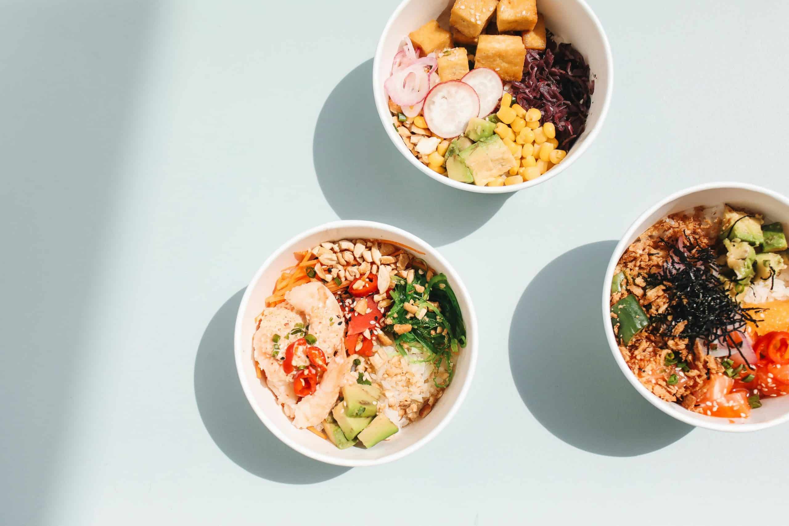 A top view of three vibrant poke bowls with varied fresh ingredients on a light blue surface.