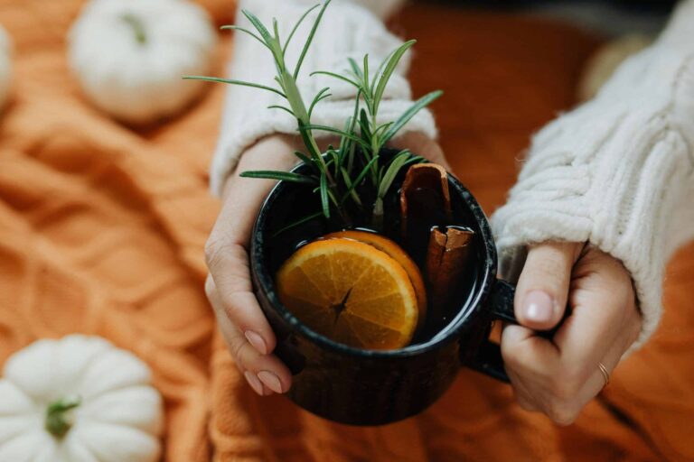pumpkin spice, Hands holding a warm cup with spices and orange slices, surrounded by autumn decorations.