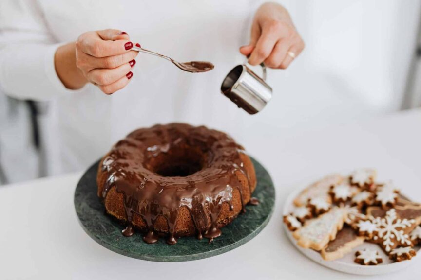 Close-up of hands adding chocolate glaze to a Bundt cake beside festive cookies.