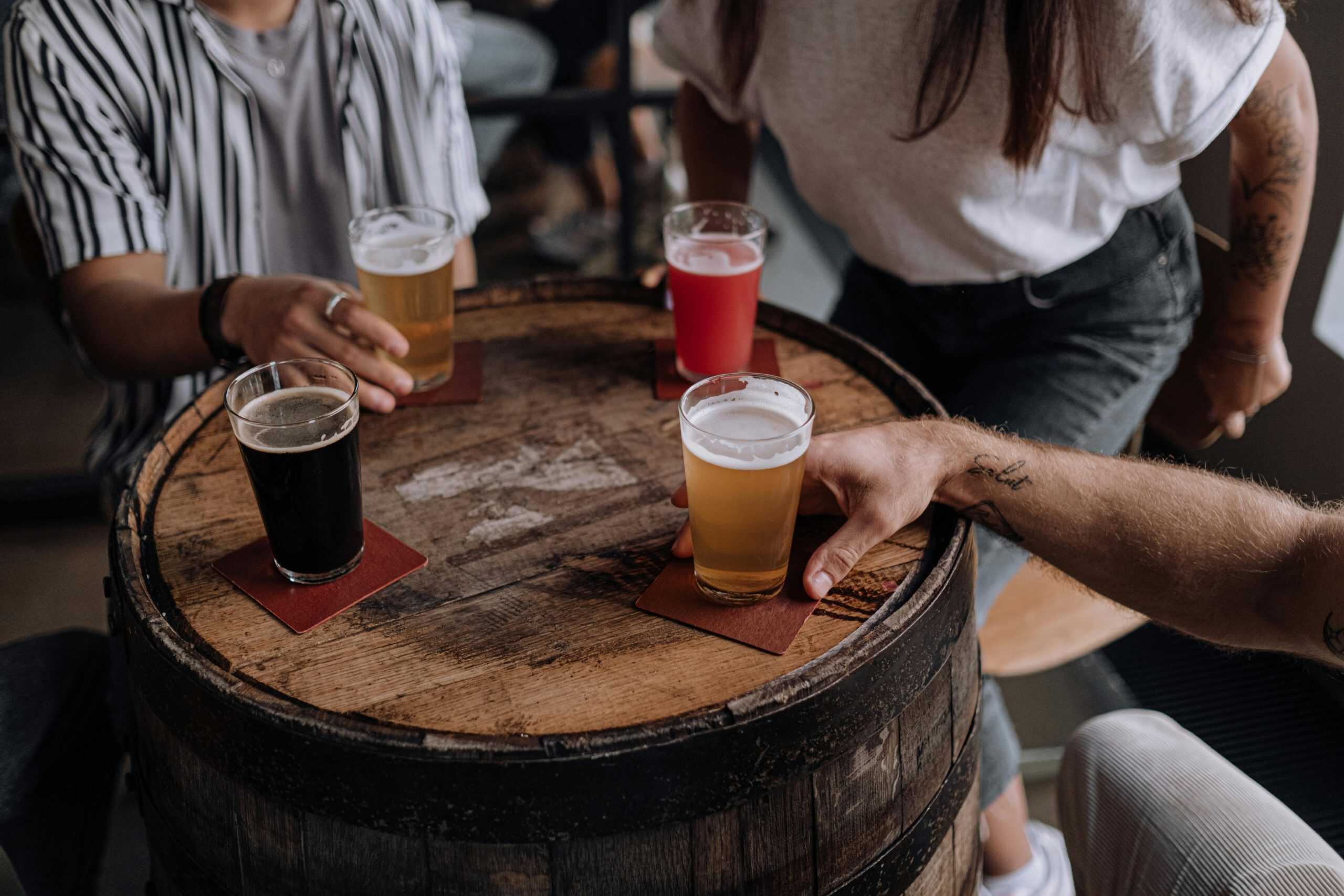 Friends enjoying craft beers around a wooden barrel table in a cozy indoor setting.
