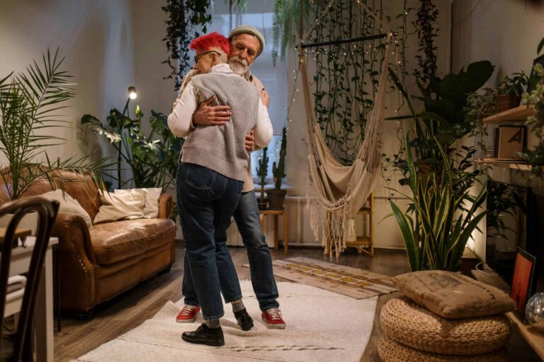 An elderly couple shares a tender embrace in a tranquil, plant-filled living room setting. accessibility