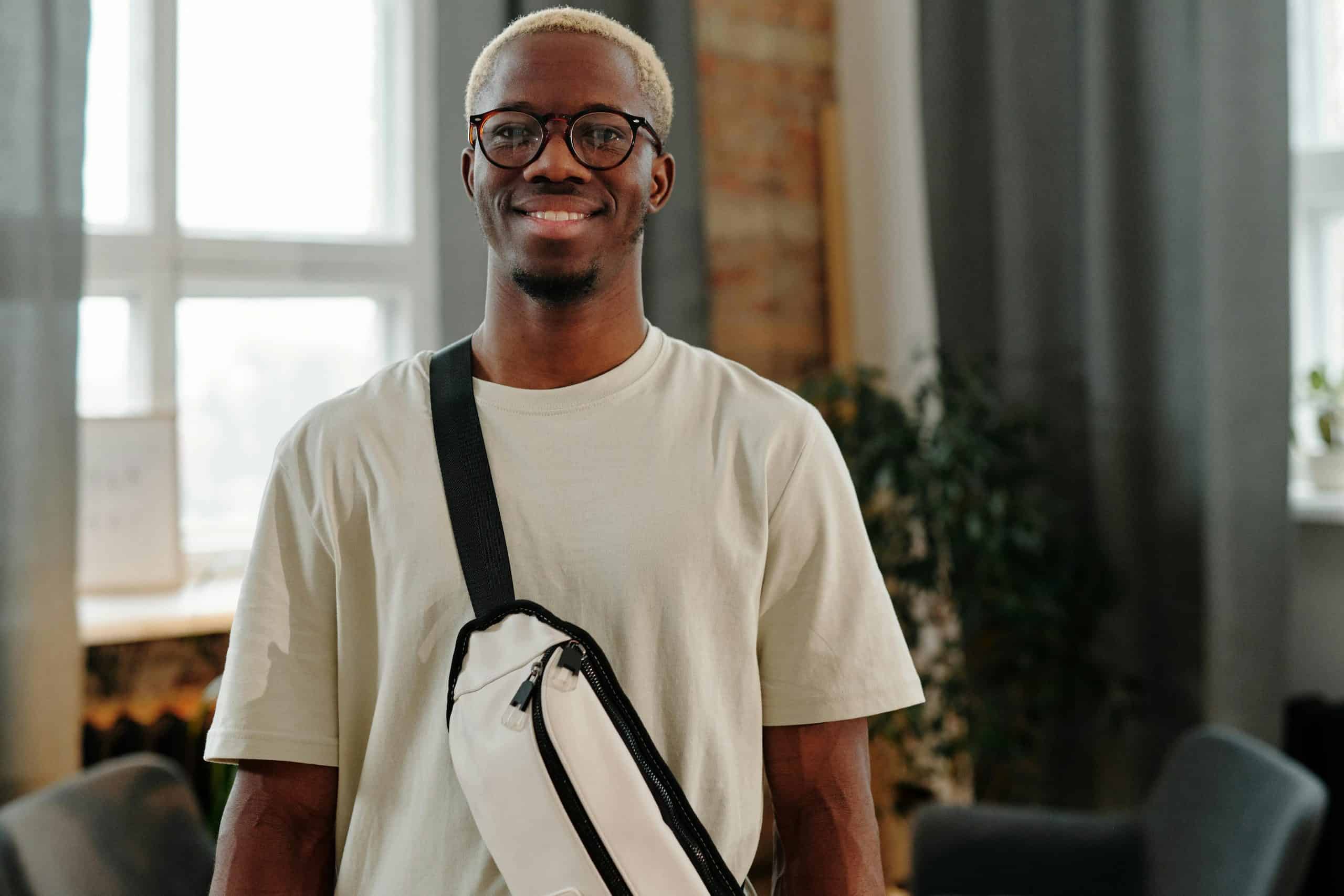 Casually dressed man with eyeglasses and bag smiling indoors against a bright window.