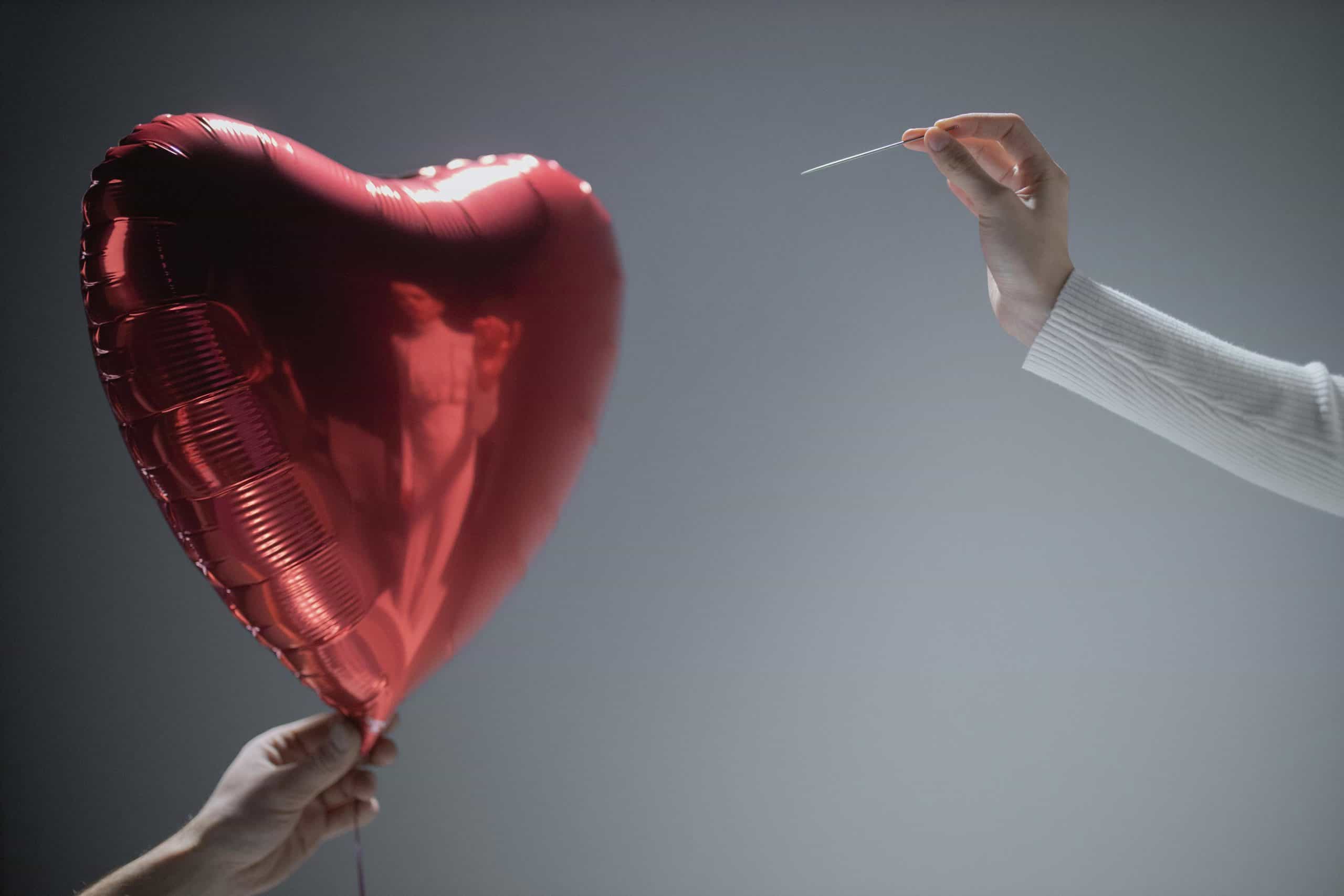 Close-up of a red heart-shaped balloon being popped by a needle, evoking themes of love and fragility.