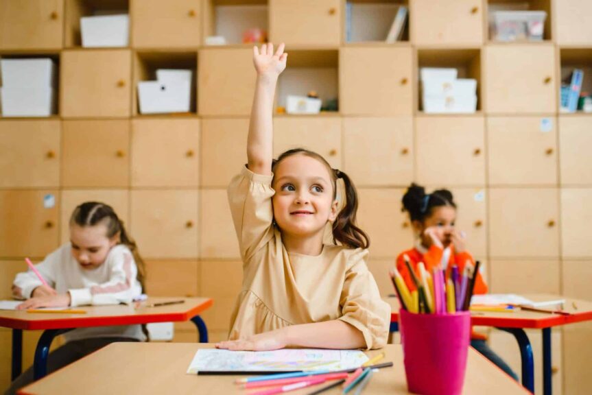 Three children engaged in learning activities inside a classroom, with one raising her hand, back-to-school, immunizations
