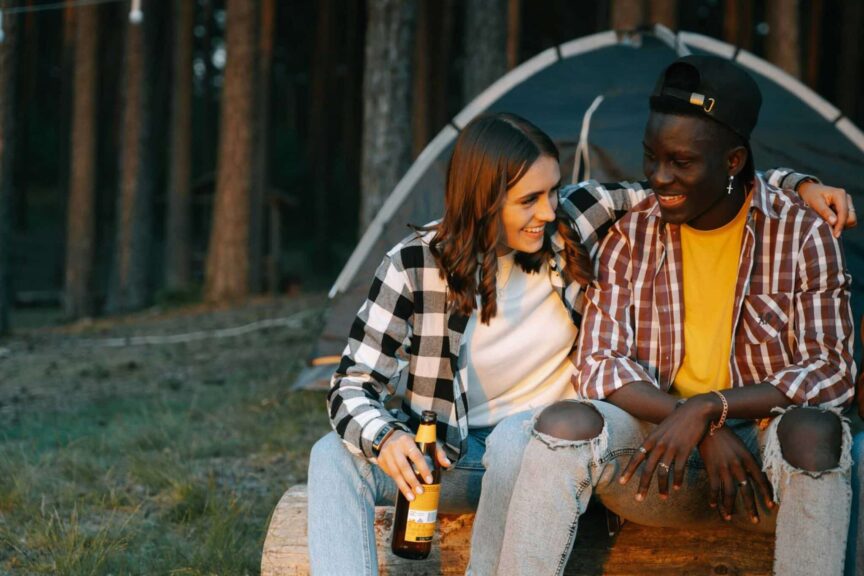 Two friends enjoying a cheerful outdoor camping trip, sitting on a log, smiling with beer bottles. Fall camping, cool-weather