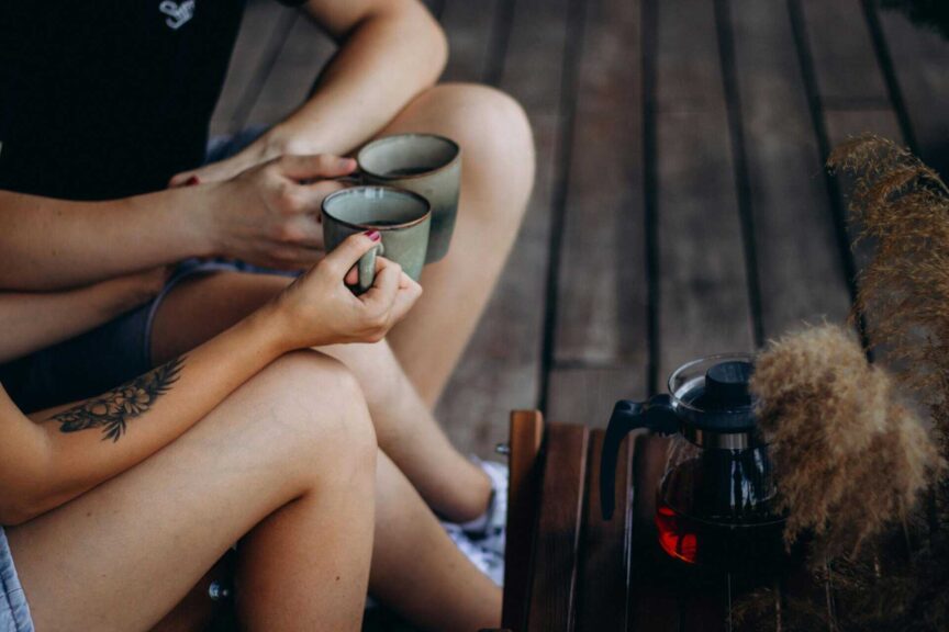 Two adults holding coffee cups, sitting together with visible tattoos, creating a cozy atmosphere, relationship patterns, fall