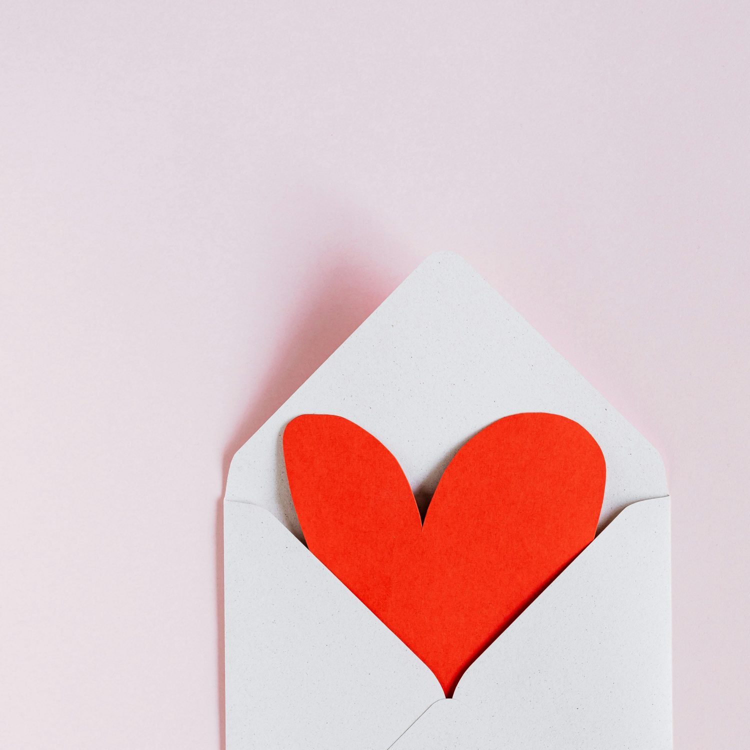 Top view of opened white envelope with handmade red paper heart placed against pink background.