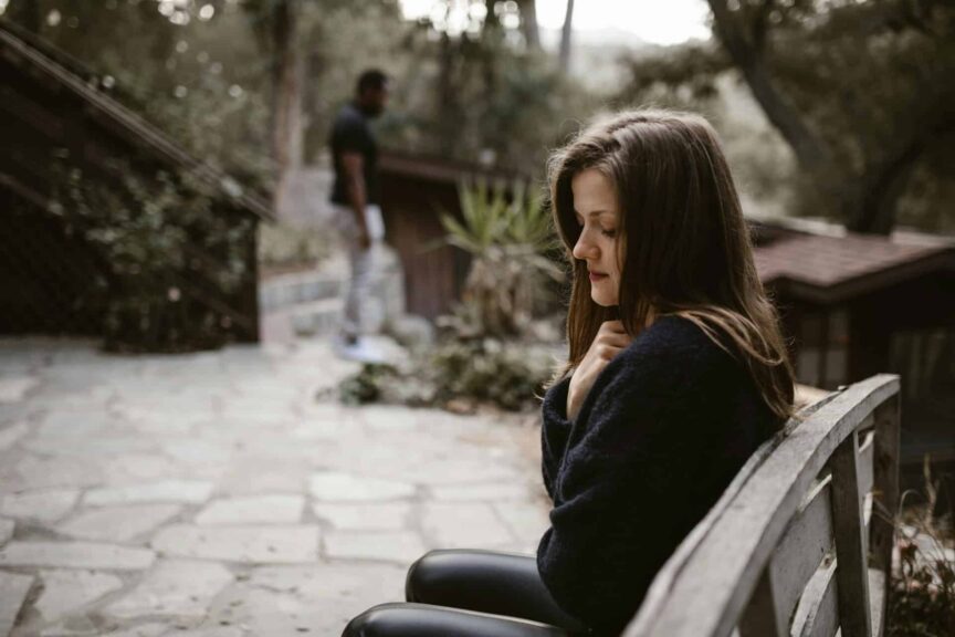A woman sitting alone on a bench with a distant person, evoking a contemplative mood. break up, loneliness