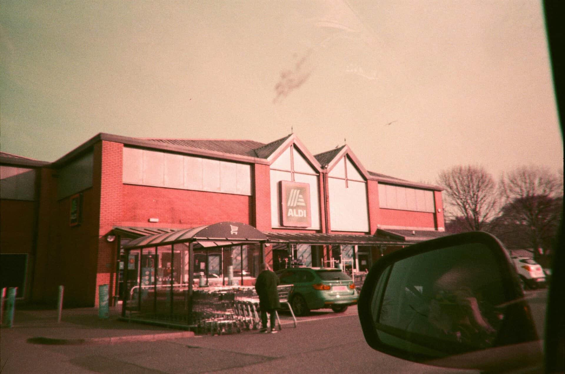 Exterior of Aldi supermarket with cars and side mirror reflection in the foreground.