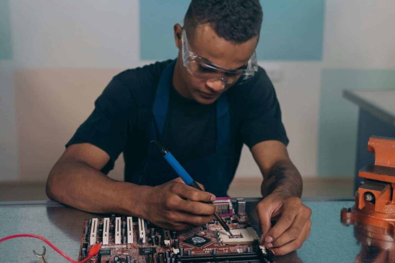 A technician wearing safety glasses works intently on a computer motherboard with a soldering iron. AI Data Centers