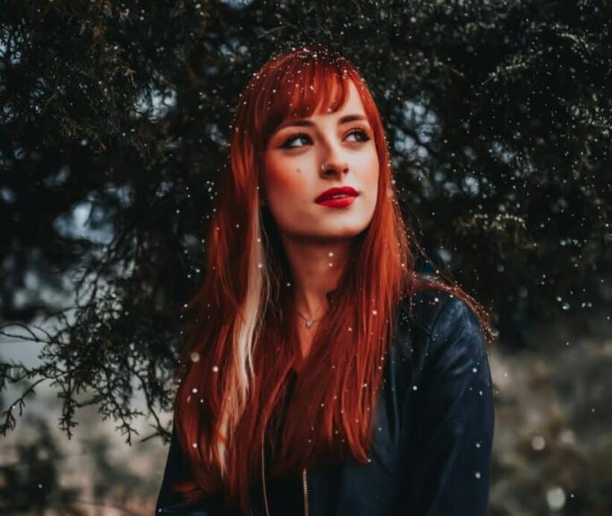 A captivating portrait of a redhead woman standing outdoors among snowflakes, wearing a black jacket. cold weather beauty mistakes to avoid
