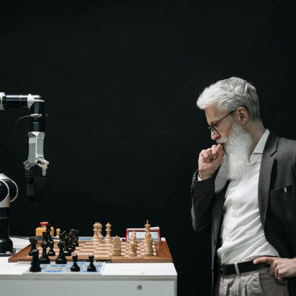 An elderly scientist contemplates a chess move against a robotic arm on a chessboard. US Government/Artificial Intelligence