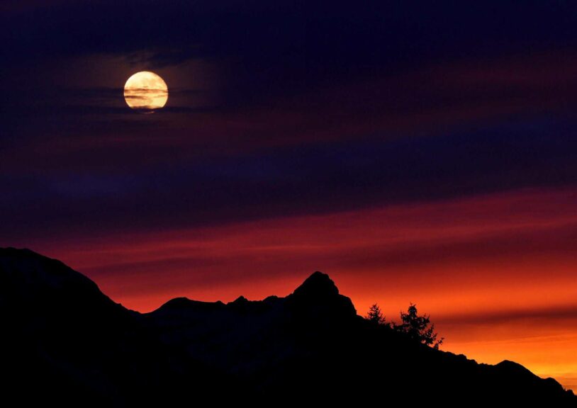 A captivating scene of a full moon above silhouetted mountain peaks against a vivid sunset sky. manifesting, Taurus Full Moon