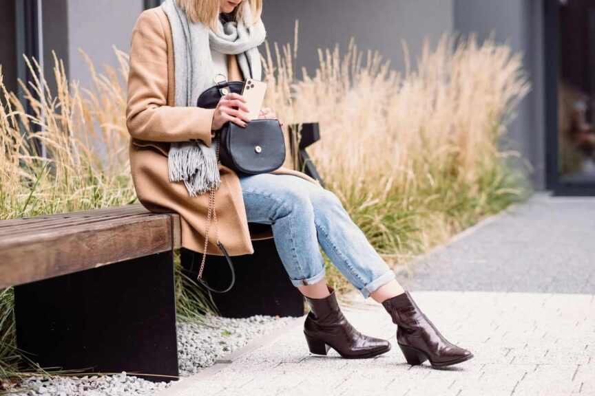 Elegant woman in a coat and scarf using her phone on a bench outdoors in Warsaw. fall boots