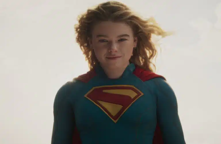 Supergirl played by Milly Alcock, wearing a blue superhero suit with a red and yellow "S" emblem, stands against a bright, cloudy sky. The mood is empowering.