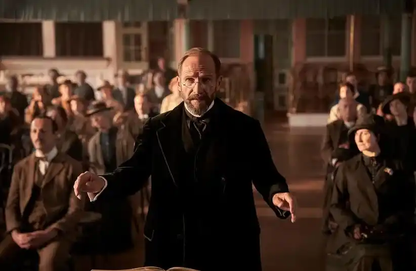 Ralph Fiennes in "The Choral" standing with a crowd of people behind him.