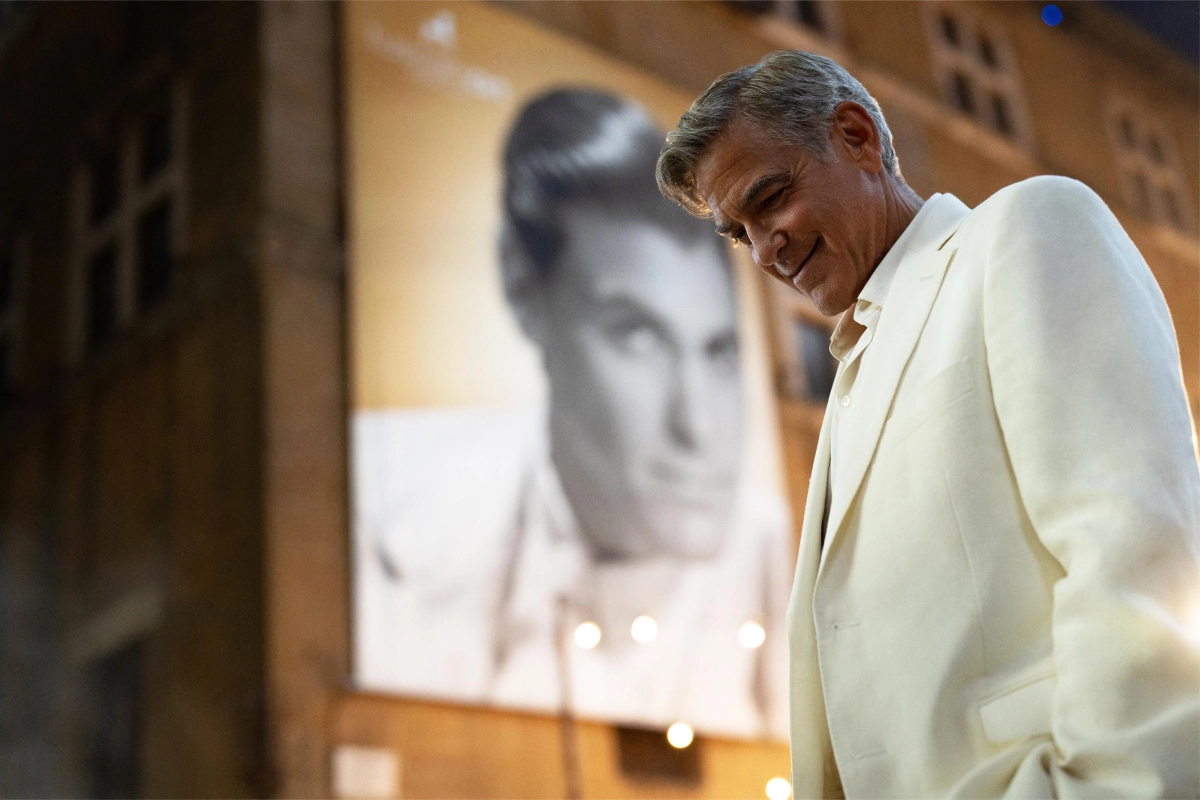 A man in a white suit is seen smiling and looking downwards. Behind him, there's a large vintage-style poster of another man. The atmosphere is warm and nostalgic.