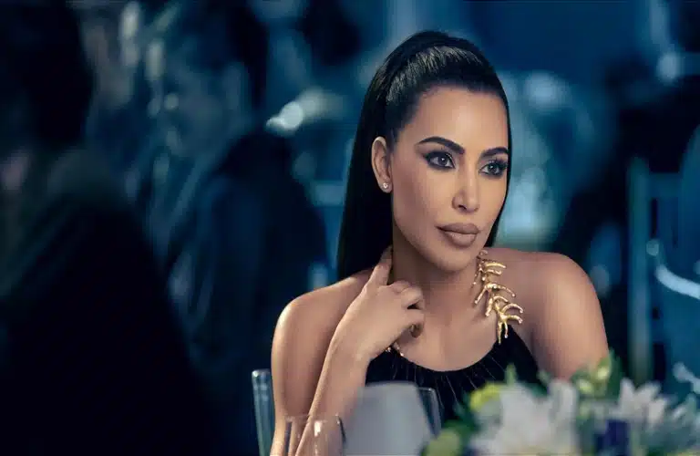 Kim Kardashian in American Horror Story (2011), with a sleek ponytail and black dress, sits thoughtfully at a dimly lit, elegant event. The gold leaf detail on her dress adds a touch of sophistication. PETA