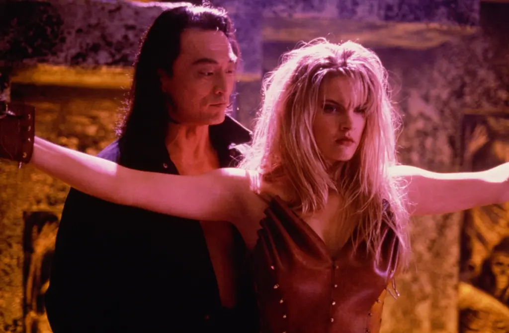 Bridgette Wilson-Sampras in Mortal Kombat (1995) stands with arms outstretched, dressed in a brown leather outfit. Cary-Hiroyuki Tagawa stands close behind her, looking intently. The setting is dimly lit, creating a tense and dramatic mood.