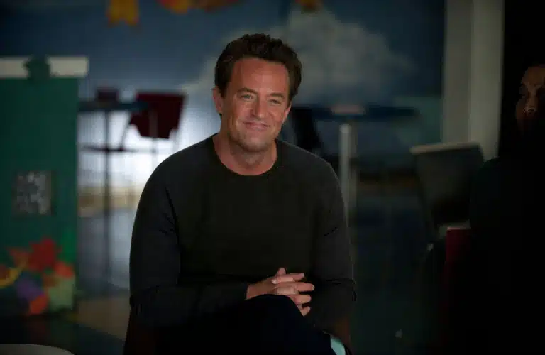 Matthew Perry smiles warmly, sitting indoors with legs crossed. The background includes colorful art and chairs, suggesting a casual setting.