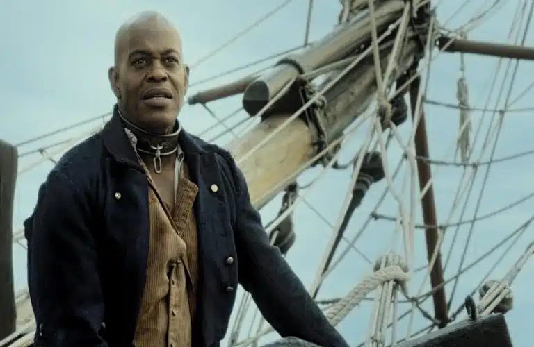 Jubilant Sykes in historical attire stands confidently on a ship's deck, with ropes and wooden masts in the background against a clear sky. The mood is determined.